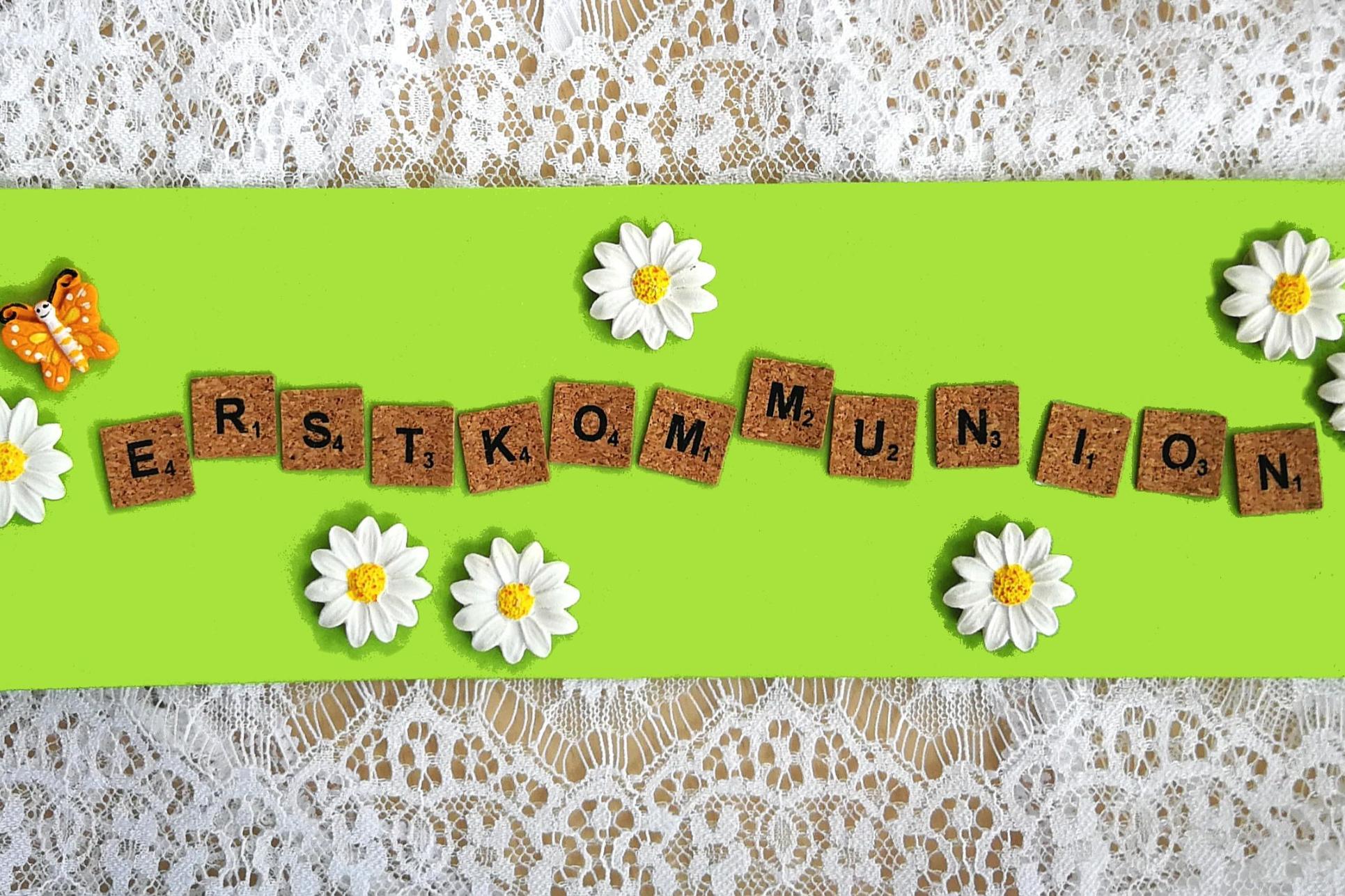 Holzbuchstaben „Erstkommunion“ auf grünem Hintergrund, umgeben von Gänseblümchen und Schmetterling.